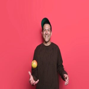 Man laughing whilst throwing an apple in the air with a pink background