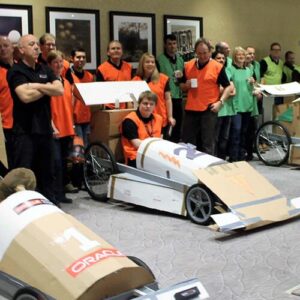 Diverse teams with a representative member sitting in a cardboard cutout race car in a room