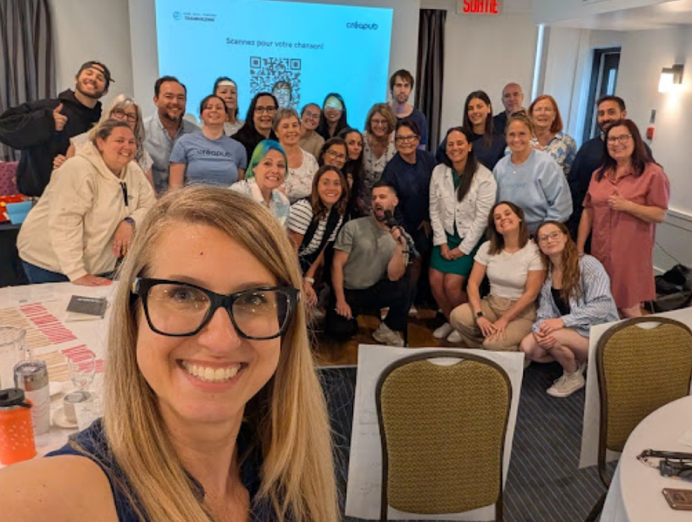Group of diverse team members smiling together at a leadership workshop celebrating Creapub’s 50th anniversary, with a screen in the background showing a QR code for their team song.