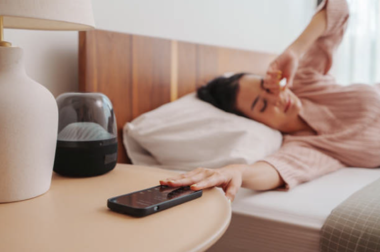 Une femme allongée dans son lit qui tend la main vers son téléphone intelligent sur la table de chevet, représentant l’habitude de consulter son cellulaire dès le réveil et comment cela affecte la concentration et le succès.