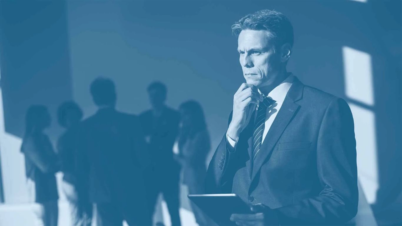 Thoughtful business leader holding a tablet while reflecting under pressure, with team blurred in the background.