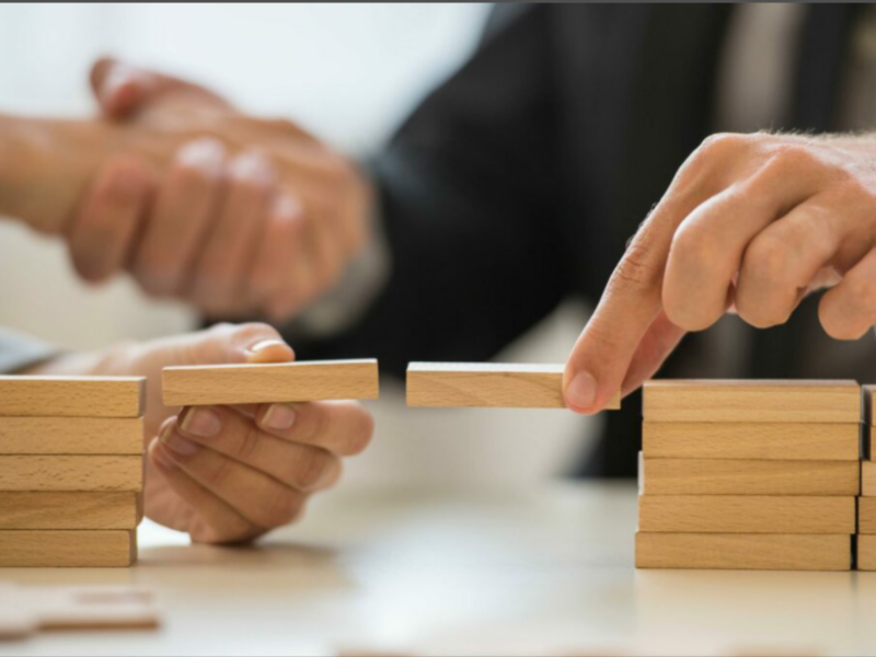 Des mains placent délicatement un bloc de bois entre deux piles, symbolisant la confiance, la sécurité psychologique et le lien créé par la présence du leader.